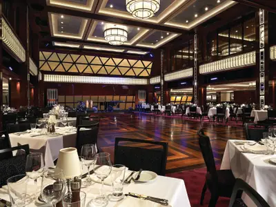 Ein weitläufiges, elegantes Restaurant an Bord der Norwegian Getaway. Der Raum verfügt über eine große Tanzfläche, eine Bühne mit Instrumenten und zahlreiche weiß gedeckte Tische, umgeben von dunklem Holz und stilvoller Beleuchtung. Die Atmosphäre ist einladend und bereit für Gäste.