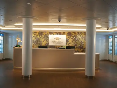 Mandara Spa & Salon Empfangsbereich an Bord der Norwegian Getaway. Heller Tresen, zwei Säulen, goldene Mosaikwand mit Logo, seitlich beleuchtete Produktvitrinen.