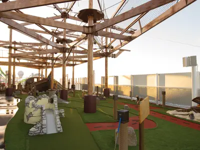 Mini-Golf und Hochseilgarten auf der Norwegian Getaway bei Sonnenuntergang. Grüne Bahnen, Schiffswrack und Holzkonstruktion des Klettergartens sind zu sehen.