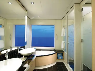 Geräumiges Badezimmer an Bord der Norwegian Getaway. Es zeigt eine Badewanne mit Meerblick, zwei Waschbecken, eine separate Duschkabine und frische Handtücher.