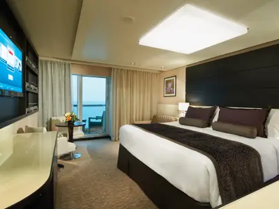Eine elegante Suite an Bord der Norwegian Getaway mit großem Bett, TV, Sitzbereich und Balkon mit Meerblick. Braune und beige Farbtöne dominieren den Raum.