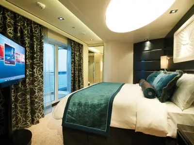 Eine luxuriöse Suite mit großem Bett, Fernseher und privatem Balkon mit Meerblick an Bord der Norwegian Getaway. Moderne Einrichtung mit dunklen Akzenten und tealgrünen Kissen.