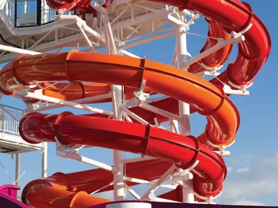 Zwei große, bunte Wasserrutschen, eine orange und eine rot, winden sich um weiße Stützstrukturen unter strahlend blauem Himmel an Bord der Norwegian Getaway.