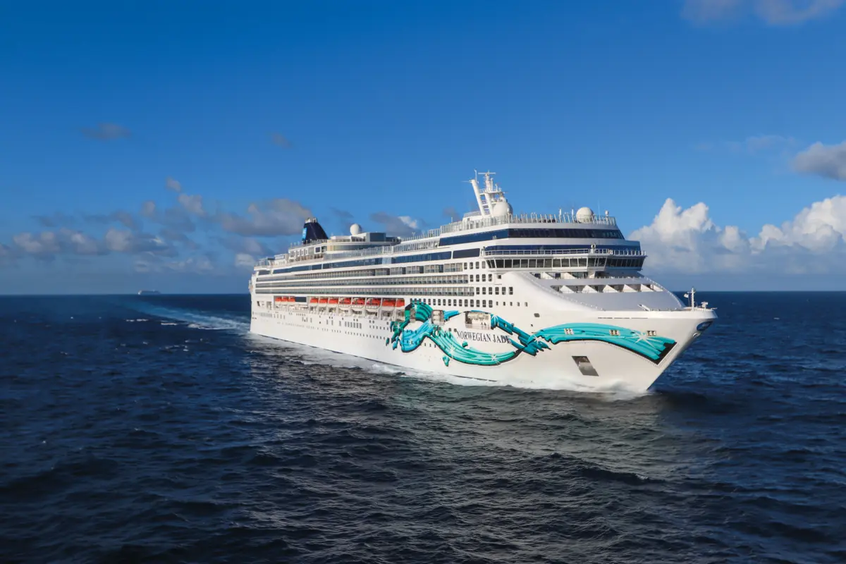 Die Norwegian Jade, ein weißes Kreuzfahrtschiff mit markantem türkisfarbenem Design, fährt auf dem offenen Meer. Blauer Himmel mit Wolken, ein weiteres Schiff am Horizont.