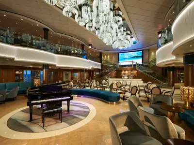 Blick auf das grosszuegige, modern gestaltete Atrium der Norwegian Jade mit Bar, Flügel, komfortablen Sitzgruppen und Videowand. Eine grosse Videowand zeigt eine tropische Strandlandschaft.