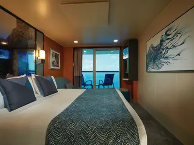 Eine moderne Balkonkabine an Bord der Norwegian Jade. Im Vordergrund ein grosses Doppelbett mit blauer Tagesdecke. Dahinter Balkontür mit Meerblick und Stühlen. Kunst an den Wänden.