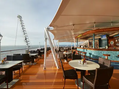Weitwinkelaufnahme der Great Outdoors Bar mit Tischen, Stühlen und einem Dach auf dem Deck der Norwegian Jade. Das Meer ist im Hintergrund sichtbar, der Boden glänzt im Sonnenlicht.