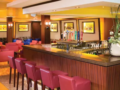 Eine leere Bar an Bord der Norwegian Jade. Eine lange Theke mit roten Barhockern, dahinter Zapfhähne und Spirituosenregale. Im Hintergrund rote und blaue Lounge-Sessel und Tische.