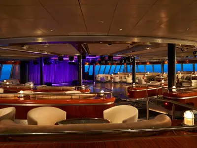Die Spinnaker Lounge an Bord der Norwegian Jade. Elegante Sitzgruppen, Holzelemente und grosse Fenster mit Meerblick. Eine Bühne mit lilafarbenem Vorhang ist im Hintergrund zu sehen.