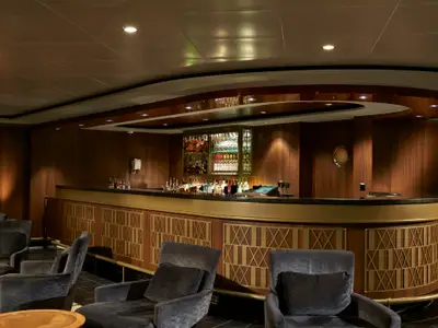 Die Sugarcane Bar an Bord der Norwegian Jade. Eine halbrunde Bar mit goldener Verkleidung und Spirituosenregal. Gemütliche Sessel in dunklem Farbton laden zum Verweilen ein. Warme Holzvertäfelung.
