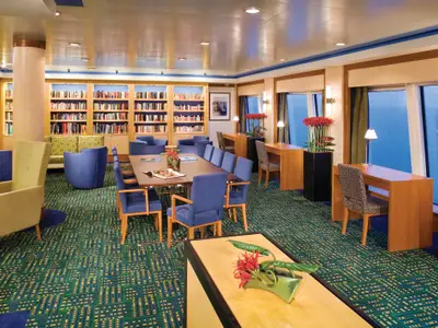 Die helle und einladende Bibliothek an Bord der Norwegian Jade. Zahlreiche Bücherregale, eine lange Lesetafel, bequeme Sessel und Tische sowie grosse Fenster mit Meerblick sind zu sehen.