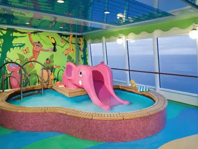 Bunter Kinderpool-Bereich an Bord der Norwegian Jade. Eine pinke Elefantenrutsche führt in ein flaches Becken. Die Wand ziert ein Dschungel-Wandbild mit Tieren. Große Fenster bieten Meerblick. Keine Personen sichtbar.