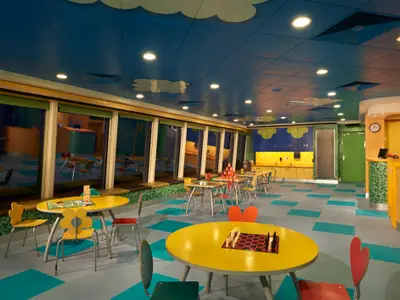 Ein lebhafter, farbenfroher Kinderbereich an Bord der Norwegian Jade mit gelben Tischen, bunten Stühlen und großen Fenstern. Spielbretter und ein Schachspiel liegen bereit.