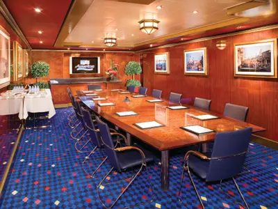 Meeting room on the Norwegian Jade with a long wooden conference table, blue chairs, a TV screen, and refreshment tables. The room features wood-paneled walls with artwork and a blue, red, and yellow checkered carpet.