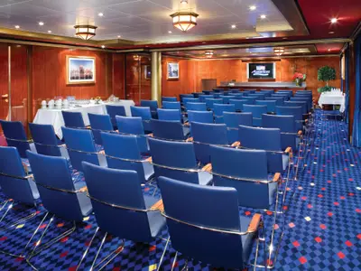 Konferenzraum an Bord der Norwegian Jade mit blauen Stühlen. Im Hintergrund stehen Buffets mit Getränken und Snacks. Eine große Leinwand ist sichtbar.