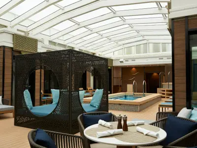 Ein heller, luxuriöser Poolbereich mit Hot Tub, dunklen Rattan-Lounges und hellblauen Kissen unter einem Glasdach an Bord der Norwegian Jade. Holz- und helle Farbtöne prägen die moderne Einrichtung.