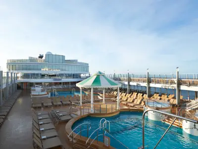 Weitläufiges Pooldeck der Norwegian Jade, umgeben von Sonnenliegen. Mehrere Pools und Whirlpools sind zu sehen, darunter ein zentraler Pool mit gestreiftem Sonnenschirm. Im Hintergrund ist das Schiffgebäude mit Glasfassade zu erkennen. Die Reling bietet einen Blick auf das Meer. Ohne Personen.