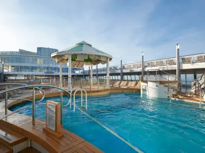 Der leere Hauptpoolbereich an Bord der Norwegian Jade unter blauem Himmel. Blaue Pools, hellbraune Liegestühle und eine runde, grün-weiß gestreifte Überdachung prägen die Szene.