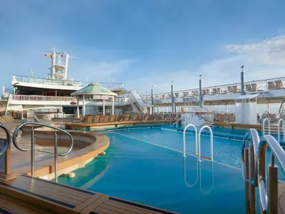 Das offene Pooldeck an Bord der Norwegian Jade, mit einem großen blauen Swimmingpool, einem Whirlpool und zahlreichen Sonnenliegen, umgeben von Schiffsaufbauten unter hellem Tageslicht. Keine Personen sind zu sehen.