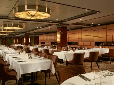 Elegantes Spezialitätenrestaurant an Bord der Norwegian Jade. Braune Ledersessel, weiss gedeckte Tische, kunstvolle Deckenleuchten und Wandpaneele mit Kamin-Elementen.