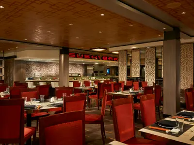 Die Sushi Bar Jasmine an Bord der Norwegian Jade zeigt Tische mit Gedecken und roten Stühlen. Im Hintergrund beleuchteter "SUSHI BAR"-Schriftzug und Theke. Modernes Interieur mit asiatischen Mustern an Decke und Wänden.