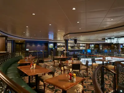 O'Sheehan's Bar & Grill an Bord der Norwegian Jade. Zweistöckiges Restaurant mit Tischen, Stühlen, Bar und eleganter Balustrade. Warme Beleuchtung, ohne Personen.