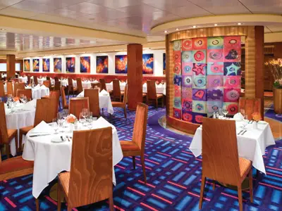 Restaurant Alizar an Bord der Norwegian Jade. Viele festlich gedeckte Tische, Holzstühle, blauer Teppich. Abstrakte bunte Kunst an Wänden und Säule.