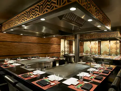 Das Teppanyaki-Restaurant Jade Teppanyaki auf der Norwegian Jade. Gedeckte Tische an Kochstationen, dunkle Stühle. Elegante Holzvertäfelung und goldene Deckenmuster schaffen ein stilvolles Ambiente.