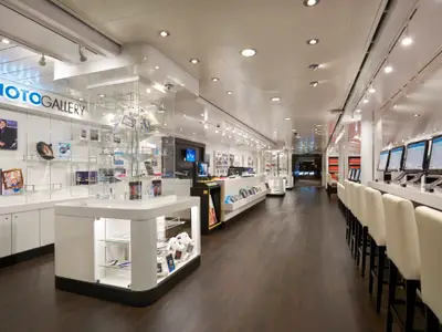 Die moderne PhotoGallery an Bord der Norwegian Jade. Helle Einrichtung mit weißen Auslagen, Computern und dunklem Holzboden. Zahlreiche Fotos sind ausgestellt.