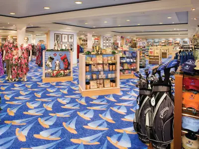 Innenansicht der Galleria Boutiques an Bord der Norwegian Jade, die eine breite Palette an Kleidung, Geschenkartikeln und Golfausrüstung präsentiert. Der blaue Teppich ist mit Segelbootmotiven verziert.
