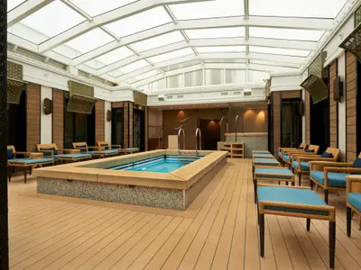 Ein heller, überdachter Poolbereich mit einem rechteckigen Becken, umgeben von warmen Holzboeden und bequemen Rattan-Sesseln mit blauen Polstern an Bord der Norwegian Jade.