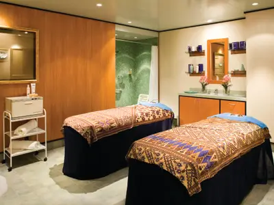 Ein geräumiger Behandlungsraum für Paare an Bord der Norwegian Jade. Er zeigt zwei Massagebetten mit gemusterten Tüchern, einen Waschtisch mit Spiegel und eine Duschkabine mit grünen Mosaikfliesen.