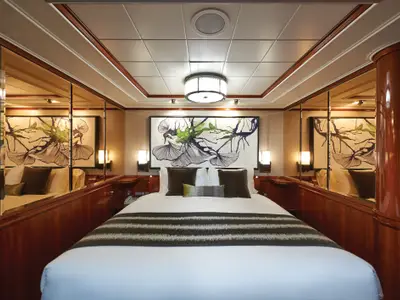 Eine elegante Suite an Bord der Norwegian Jade. Zentral steht ein großes Doppelbett mit dunklen Kissen. Ein abstraktes Kunstwerk und Holzpaneele prägen den luxuriösen Stil des Raumes.