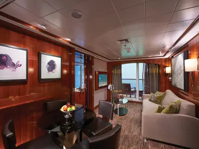 Das luxuriöse Wohnzimmer einer Suite an Bord der Norwegian Jade zeigt einen Esstisch, eine Couch, moderne Kunst an den holzvertäfelten Wänden und einen Balkon mit Meerblick.