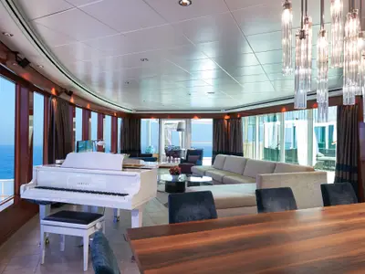 Luxuriöse Suite der Norwegian Jade. Heller Wohnbereich mit weissem Grand Piano, Eckcouch und Esstisch. Grosse Fenster mit Meerblick, ergänzt durch einen schicken Kronleuchter.