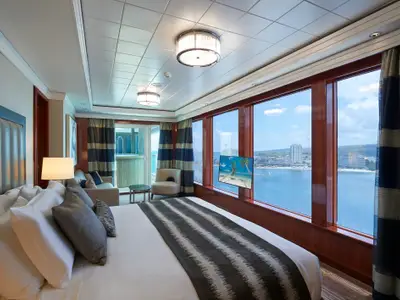 Geräumige Suite an Bord der Norwegian Jade. Heller Schlafbereich, Sitzgruppe und bodentiefe Fenster mit Panoramablick auf Meer, Strand und Küstenort bei Tageslicht.