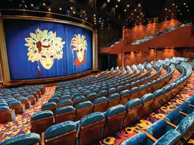 Das Stardust Theater der Norwegian Jade. Leere Bühne mit blauem Vorhang, verziert mit zwei goldenen venezianischen Masken. Blaue Samtsessel und bunter Teppich füllen den beleuchteten Theatersaal.