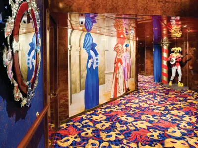 Farbenfroher Flur an Bord der Norwegian Jade. Wandmalereien kostümierter Figuren, Teppich mit venezianischen Masken. Am Gangende eine Jester-Statue mit Banjo.