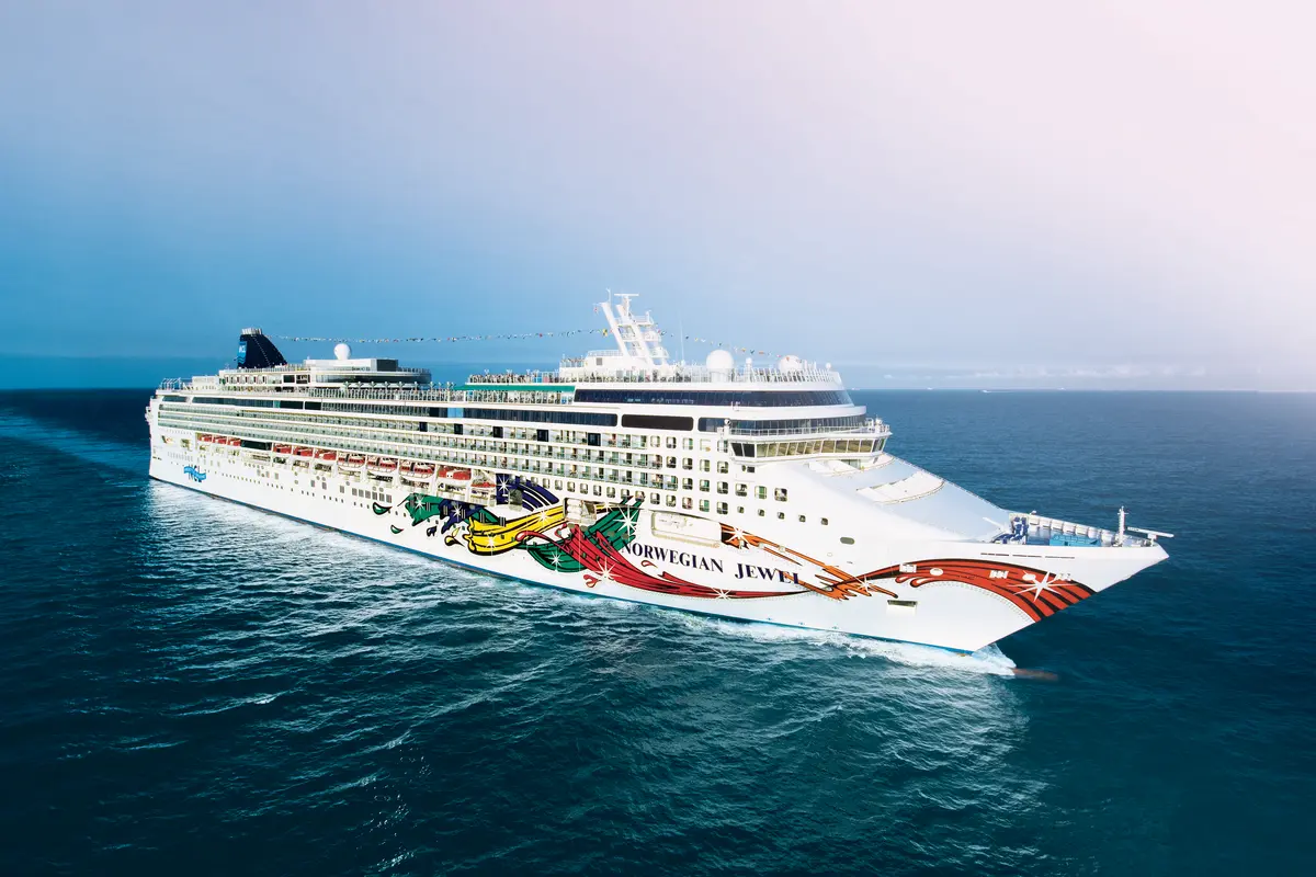 Luftbild der Norwegian Jewel, die auf dem offenen Meer fährt. Das Schiff ist von vorne rechts zu sehen, mit seiner lebhaften, künstlerischen Rumpfbemalung. Der Himmel ist oben hellblau, unten dunkler.