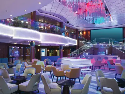 Grosses Atrium an Bord der Norwegian Jewel mit vielen Sitzgelegenheiten, einem weissen Flügel, einer Bar und breiten Treppen. Farbiges Licht erhellt den modernen Raum elegant.