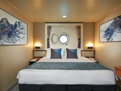 Gemütliche Aussenkabine der Norwegian Jewel mit Doppelbett, blauen Kissen, Porthole-Fenster und maritimen Kunstwerken.