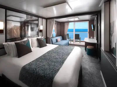 Eine helle und moderne Balkonkabine an Bord der Norwegian Jewel. Man sieht ein großes Bett, ein blaues Sofa, einen Schreibtisch und einen Balkon mit Meerblick.