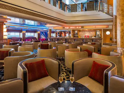 Innenansicht der luxuriösen Magnum's Champagne & Wine Bar an Bord der Norwegian Jewel mit zahlreichen bequemen Sesseln, einer gut bestückten Bar und Meerblick durch die Fenster im Hintergrund. Zwei Sektgläser im Vordergrund.
