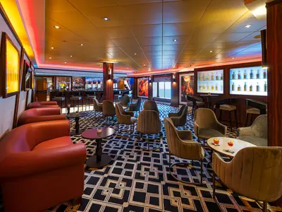 Weitwinkelaufnahme der Maltings Whiskey Bar an Bord der Norwegian Jewel. Der Raum ist mit roten Ledersofas und grauen Samtsesseln ausgestattet. Eine lange Bar, beleuchtete Flaschenregale und ein Meerblickfenster sind sichtbar.