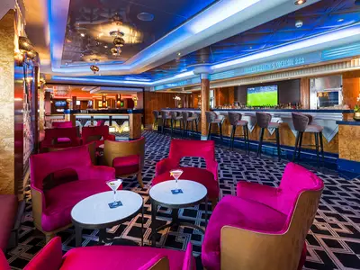 Die Shakers Martini & Cocktail Bar der Norwegian Jewel. Innenraum mit pinken Sesseln, blauer Bar, Spiegeldecke und Lichtinstallationen. Zwei Cocktails auf Tischen, TV im Hintergrund.