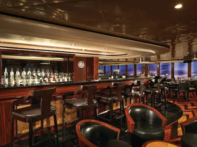Elegante Bar an Bord der Norwegian Jewel mit langer Holztheke, gefüllten Regalen und Barhockern. Große Fensterfront mit Blick auf den abendlichen Himmel und das Meer.
