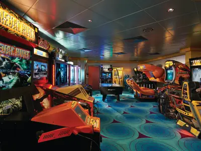 Eine beleuchtete Spielhalle an Bord der Norwegian Jewel, gefüllt mit Arcade-Automaten wie 'Primeval Hunt', 'Rambo', 'Big Buck Hunter' und 'Terminator Salvation', sowie einem Airhockey-Tisch. Der Boden hat einen gemusterten, blauen Teppich.