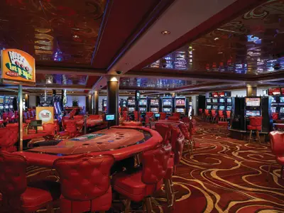Ein elegantes Casino an Bord der Norwegian Jewel mit roten Spieltischen und passenden Stühlen im Vordergrund. Im Hintergrund stehen Reihen von leuchtenden Spielautomaten. Die reflektierende Decke zeigt Muster.