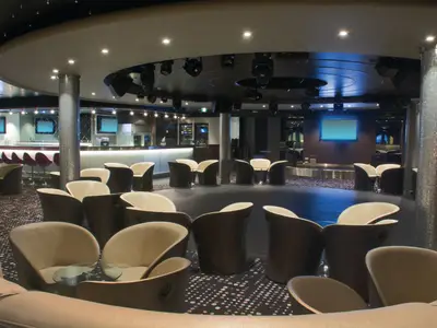 Der Nachtclub Bliss an Bord der Norwegian Jewel mit einer langen Bar, einer Bühne und zahlreichen modernen Lounge-Sesseln. Die Decke ist mit Lichtinstallationen versehen und die Säulen sind mit Mosaikfliesen bedeckt.