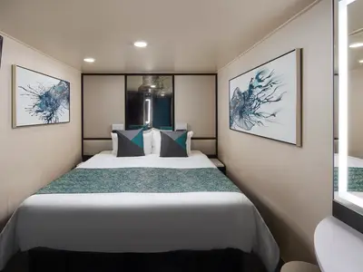 Moderne Innenkabine der Norwegian Jewel. Grosses Doppelbett mit weisser Bettwäsche und blauer Tagesdecke. Zwei abstrakte blaue Kunstwerke und ein beleuchteter Spiegel.